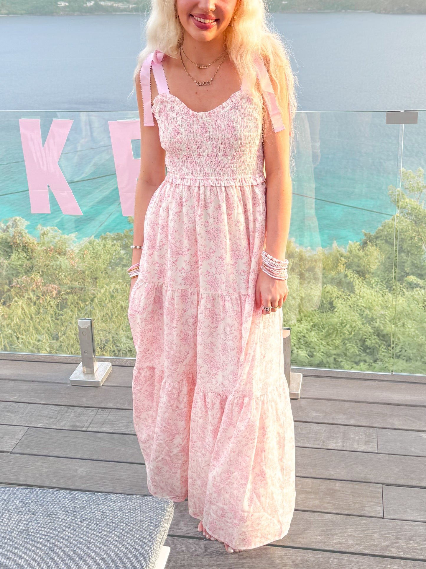 Sassy Shortcake Sweet Pea Dress — pink floral maxi with smocked bodice, tie-shoulder bows and tiered skirt on a seaside balcony.