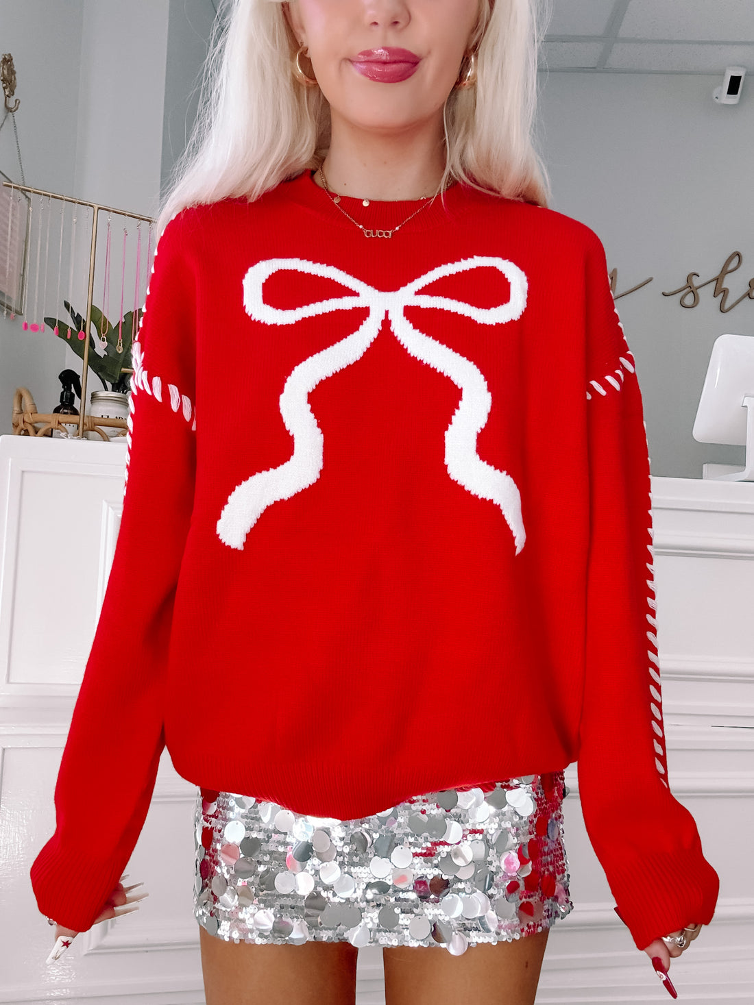 Red knit sweater featuring a large white bow design, shown styled with a silver sequined skirt.
