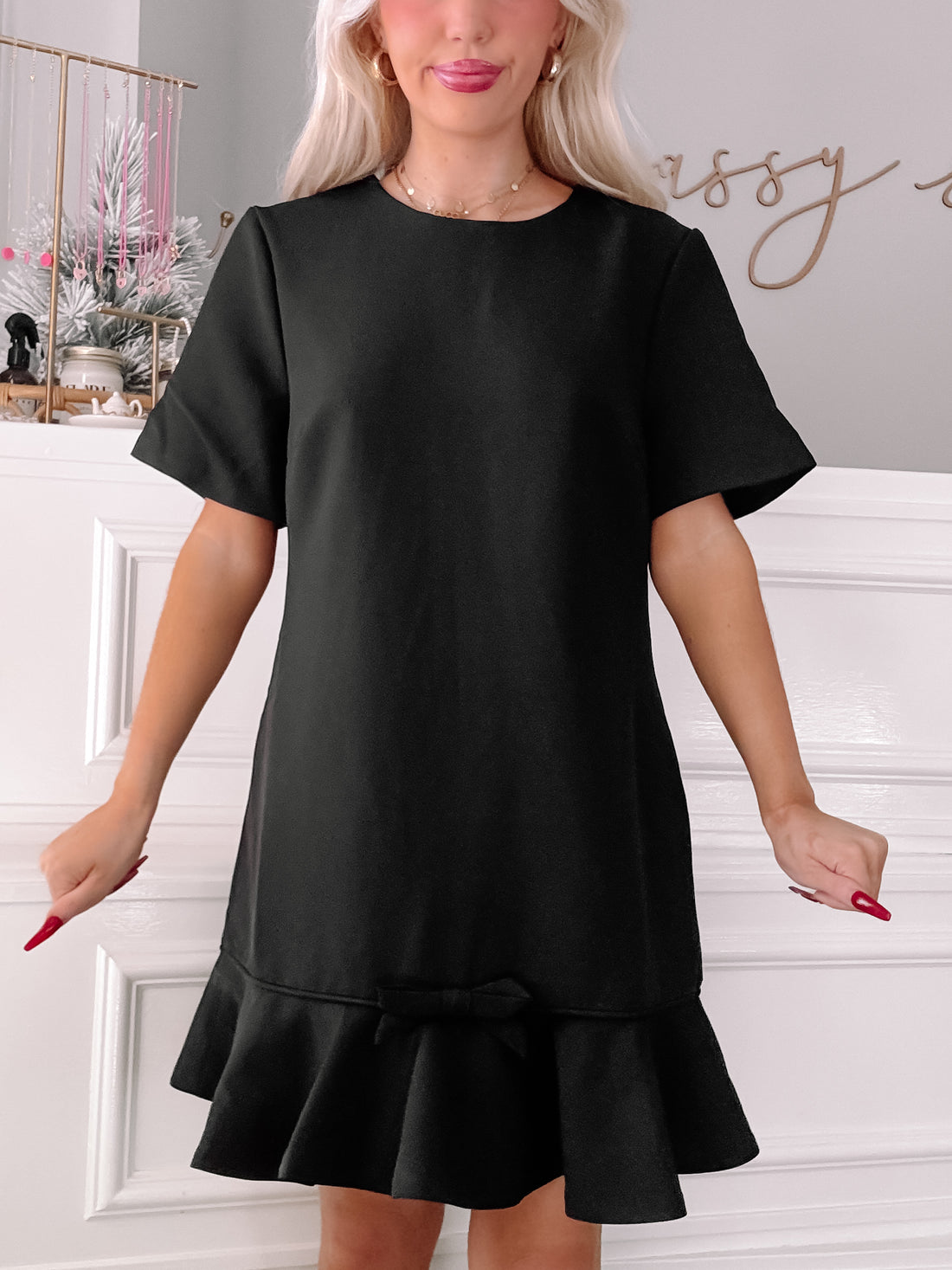 Black short-sleeve dress with a bow detail and ruffled hem, modeled indoors.
