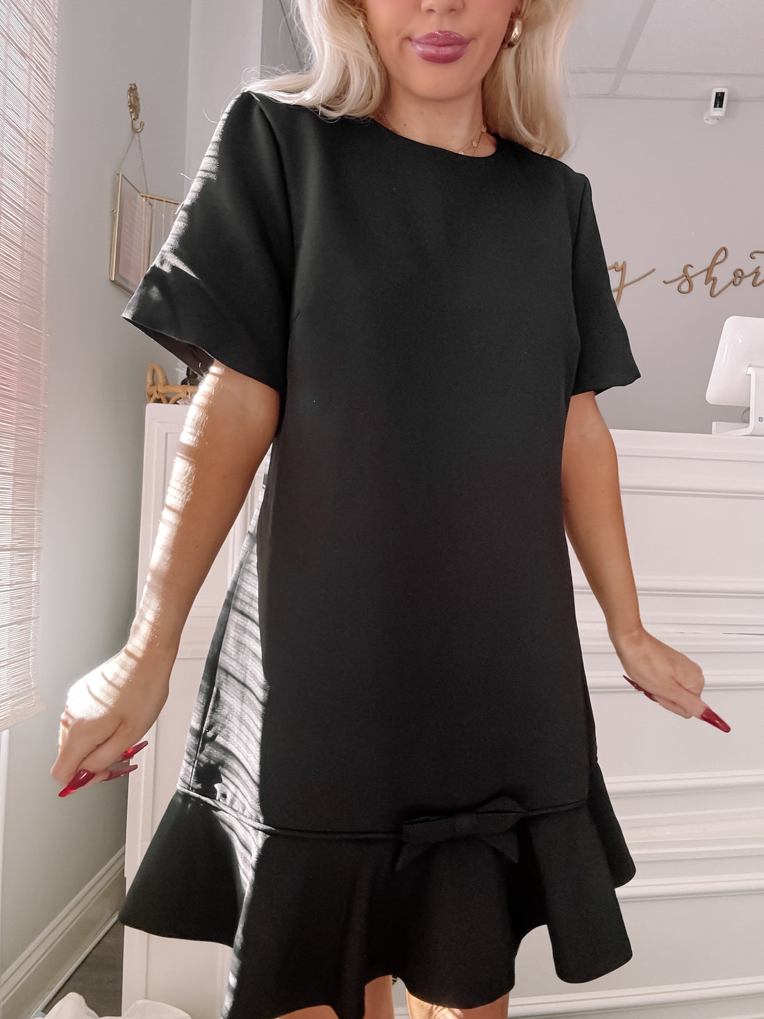 Black short-sleeved mini dress with a bow detail and ruffled hem, modeled indoors.
