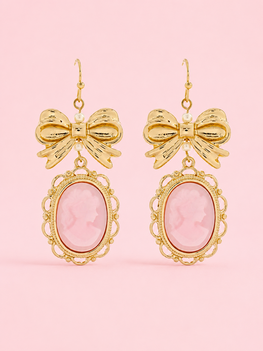 Vintage Muse Bow Earrings from Sassy Shortcake featuring gold-tone bow details, pink oval cameo charms, and fish hook backs on a light pink background.