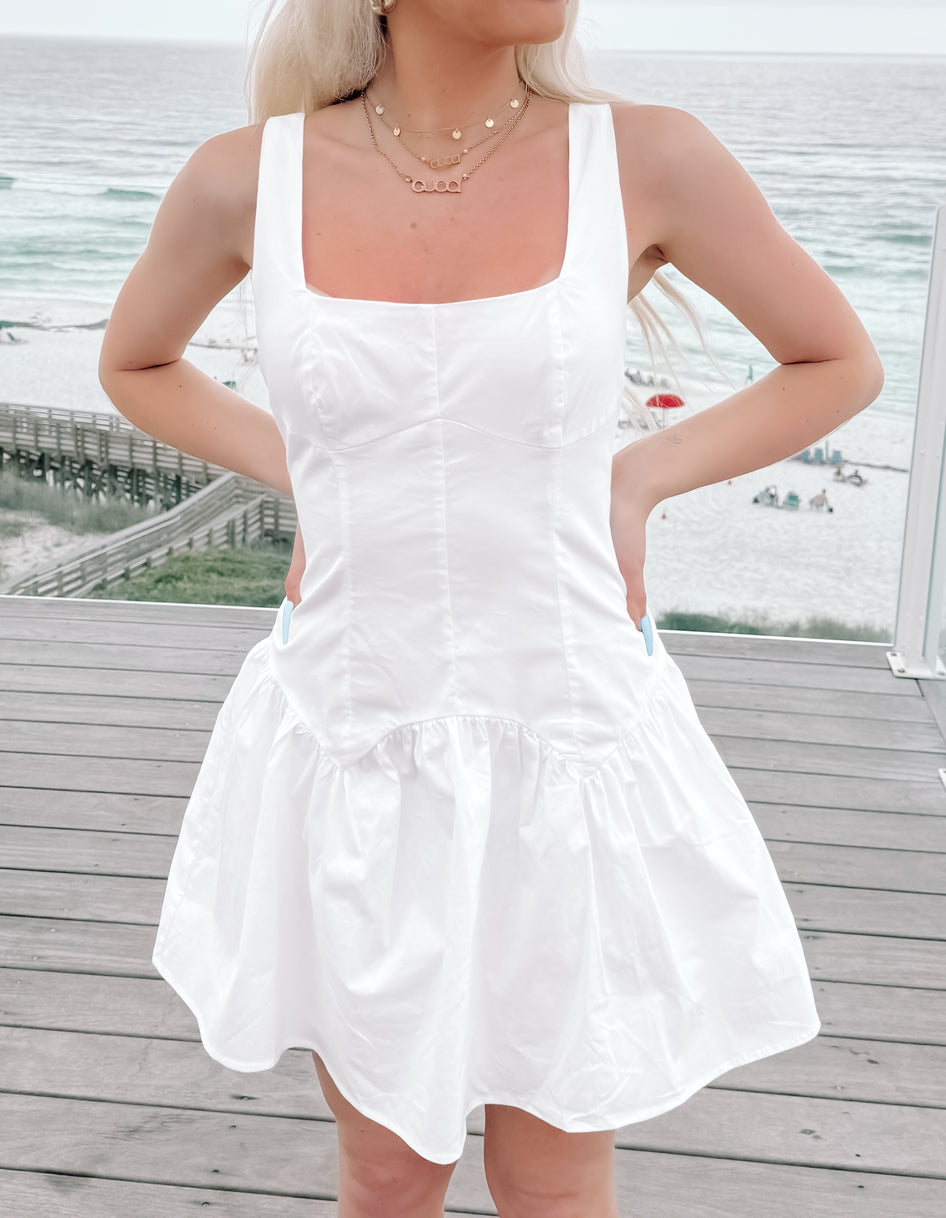White summer dress with a ruffled hem, worn outdoors near the ocean.
