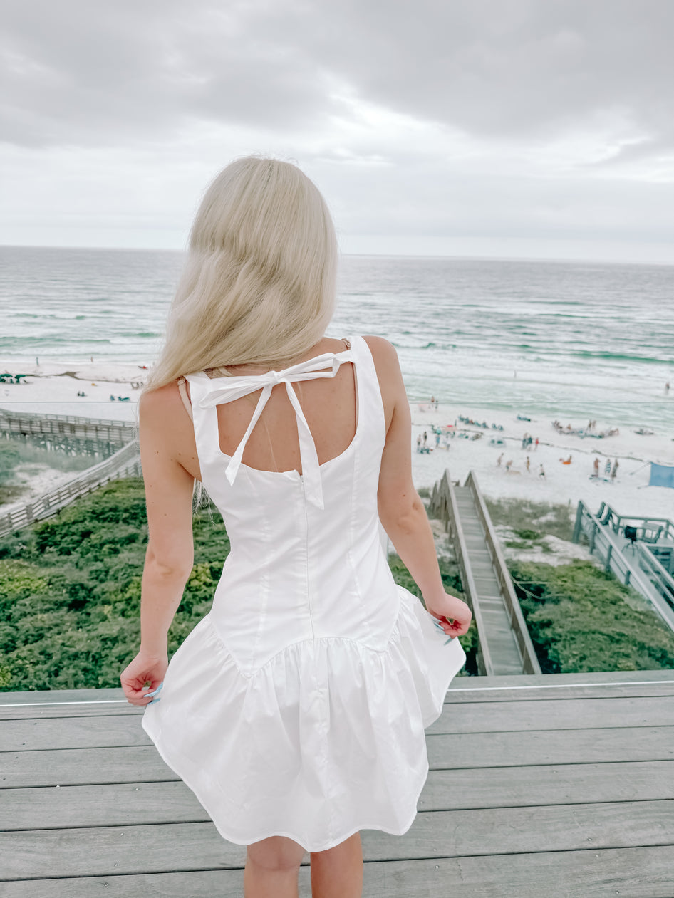 White summer dress with bow detail, viewed from behind on a deck overlooking the ocean.
