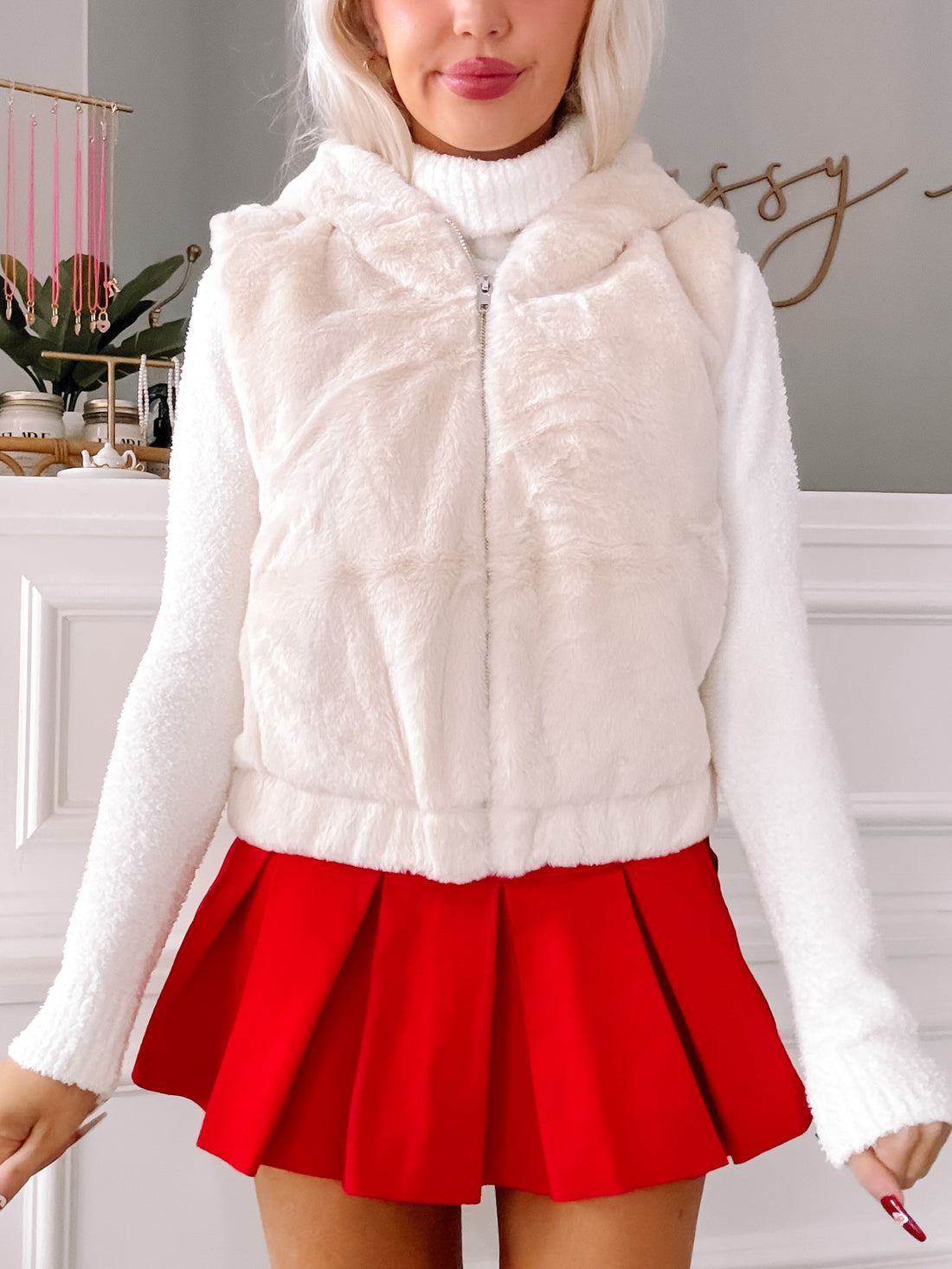 Cream-colored faux fur vest with a zipper, styled with a white sweater and red pleated mini skirt.
