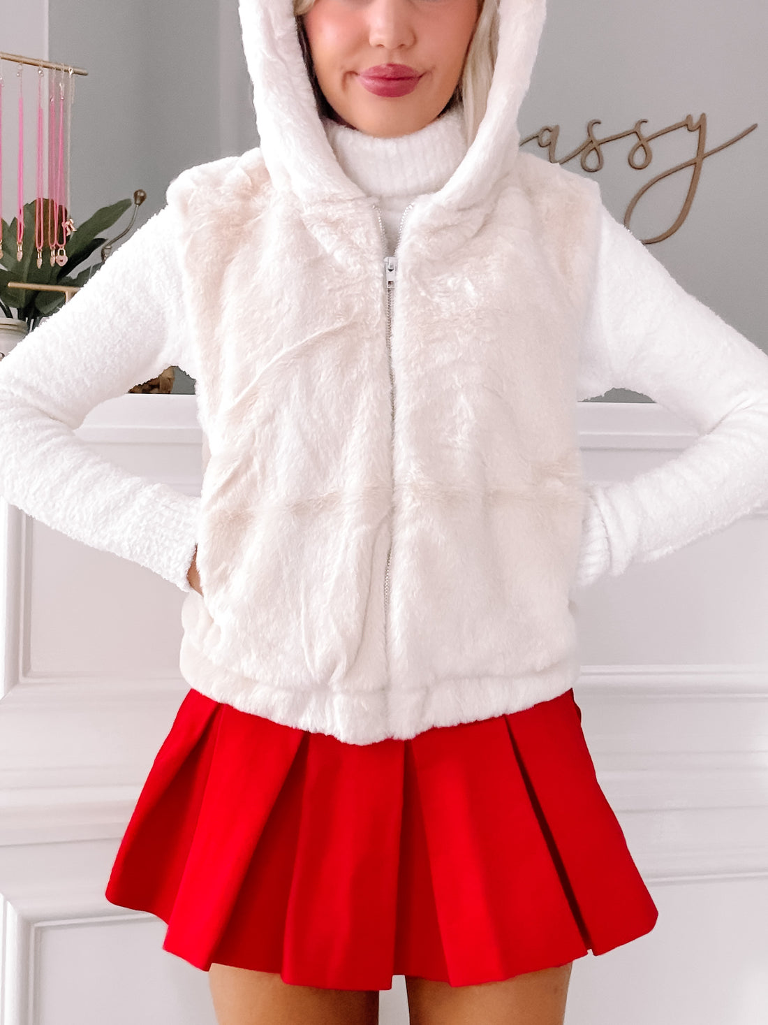 Cream faux fur zip-up vest with attached hoodie, styled with a red pleated mini skirt.
