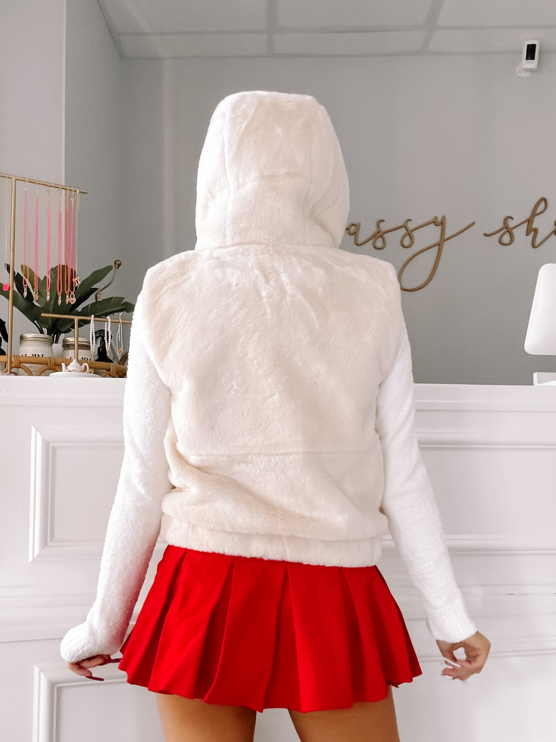 Cream-colored hooded vest with fluffy texture, worn with a red pleated skirt against a white background.
