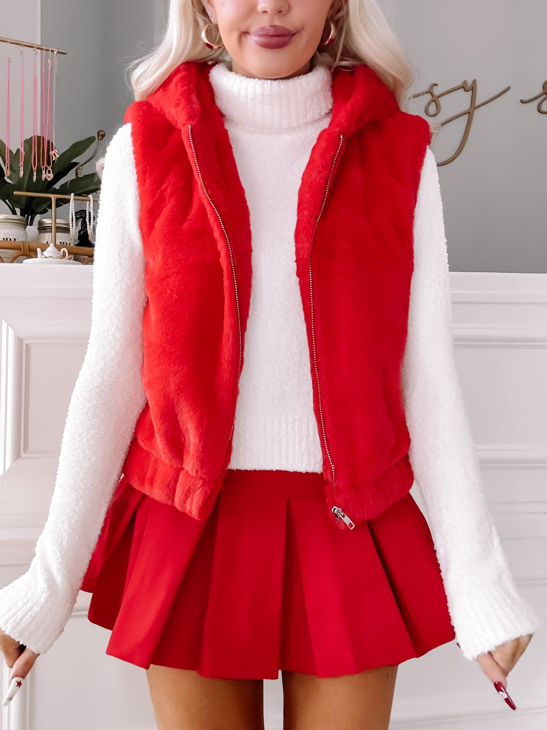 Red faux fur zip-up vest styled with a white sweater and pleated mini skirt.
