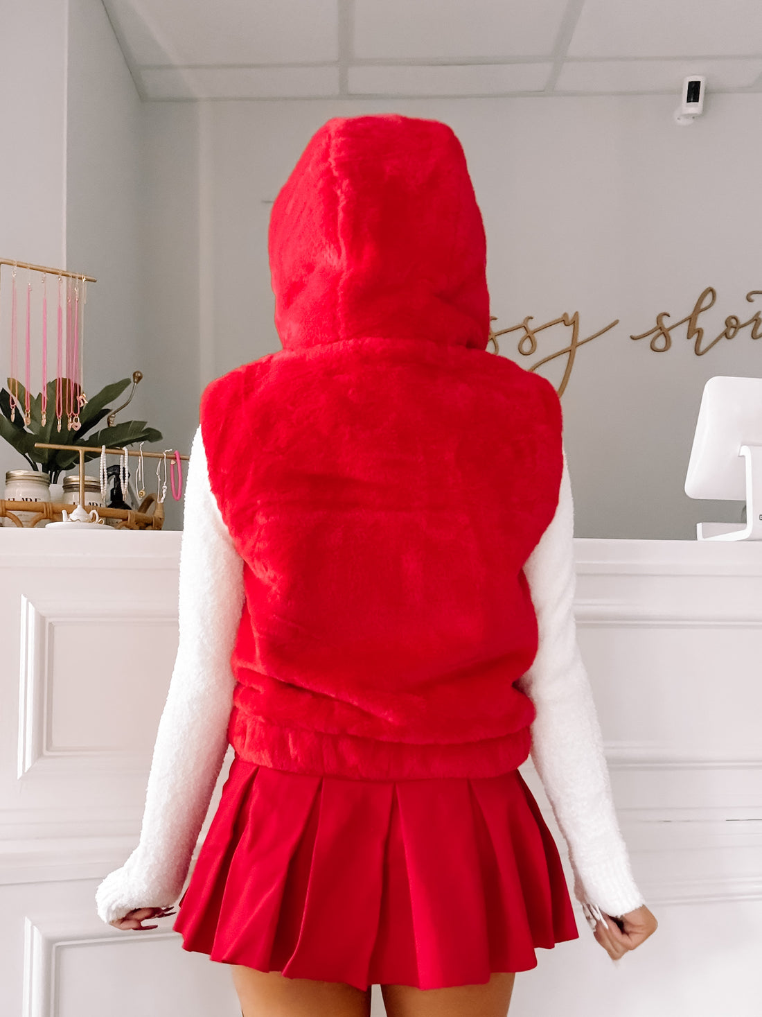 Red hooded vest with fluffy texture, paired with a white sweater and red pleated skirt.
