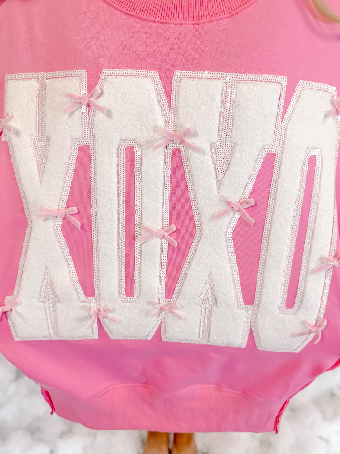 Pink crewneck sweatshirt with white XOXO lettering, accented by pink bows and sequins.
