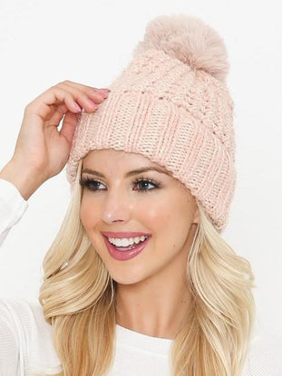 Pink knit beanie with a fluffy pompom, styled on a blonde model.
