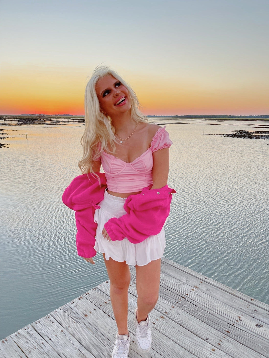 Woman in pink top and fuchsia jacket on a wooden dock at sunset.
