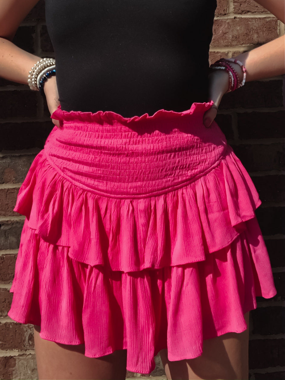 Hot pink tiered mini skirt with smocked waistband, shown against a brick wall.

