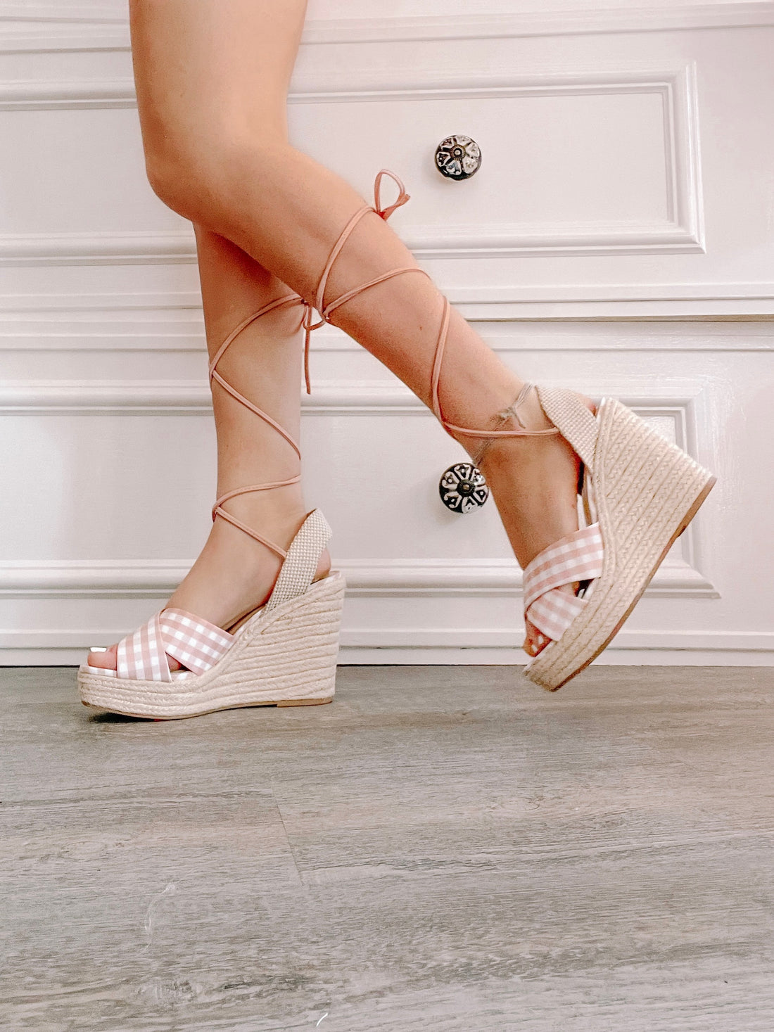 Pink gingham wedge sandals with lace-up ankles, shown on a wood floor.
