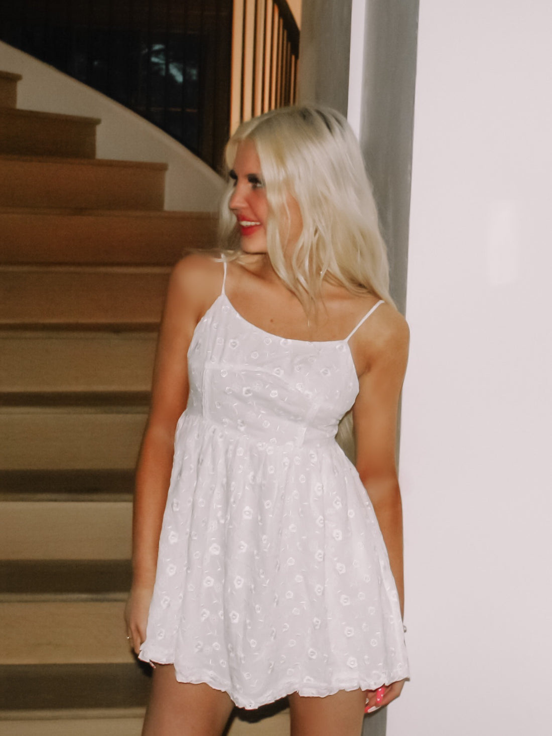 White eyelet mini dress with a fitted bodice and flared skirt, shown on a model near a staircase.
