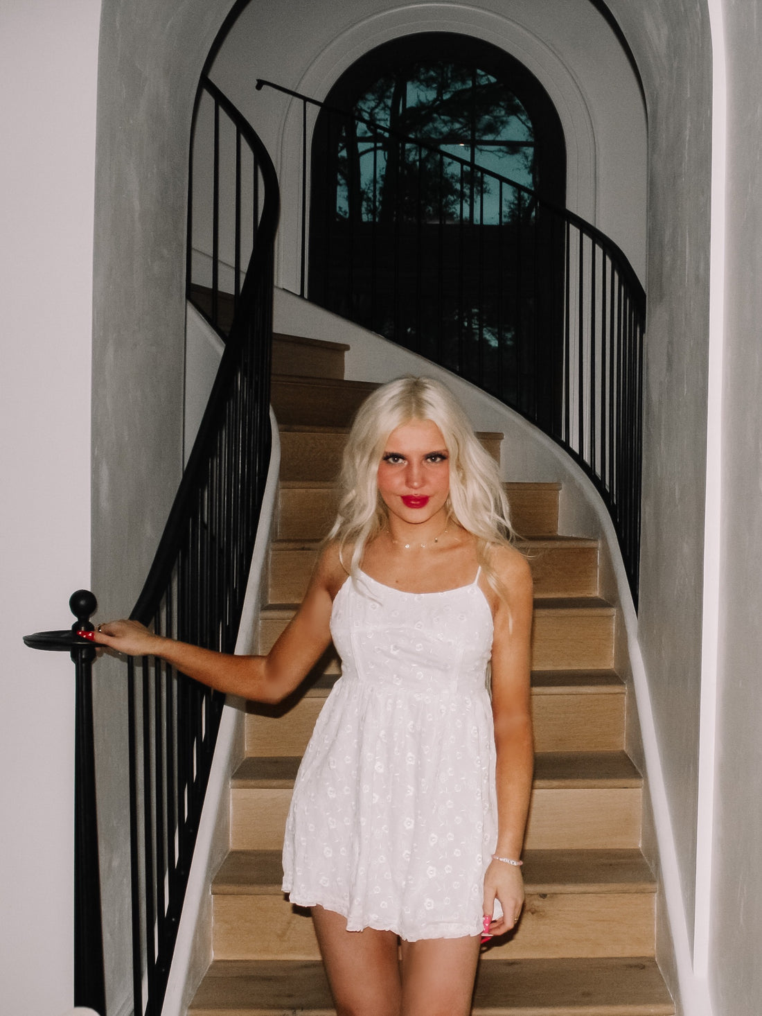 White eyelet mini dress on a blonde model, posed on a modern staircase.
