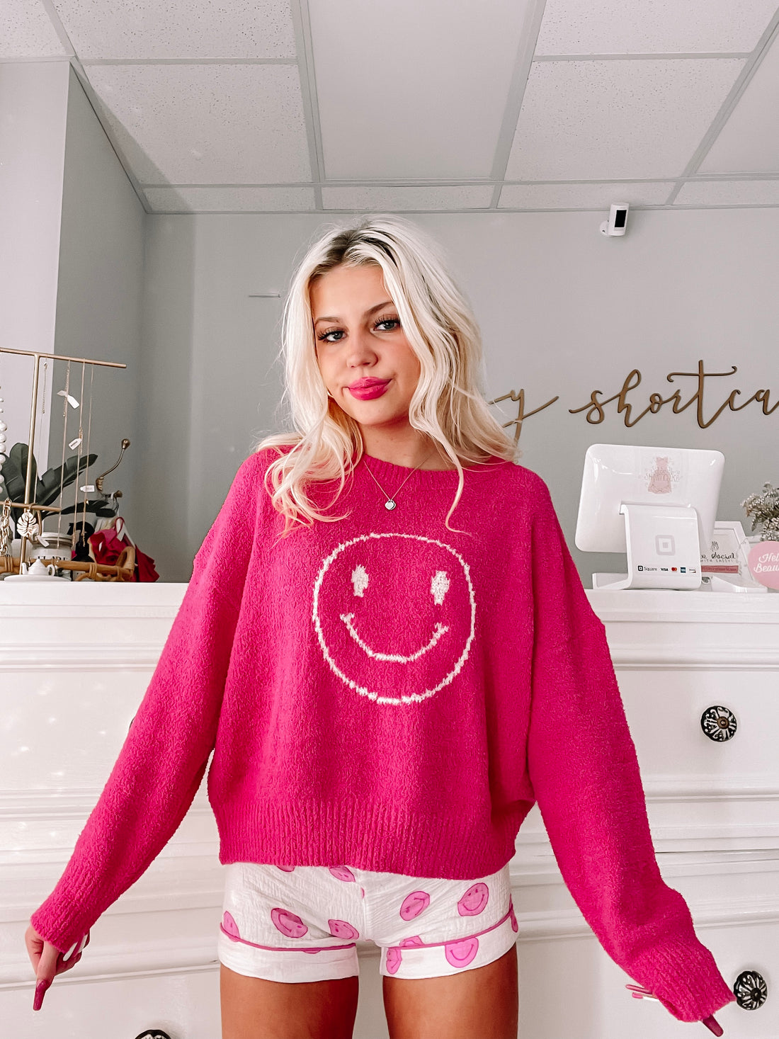 Fuzzy pink sweater and matching shorts featuring a cheerful smiley face design, modeled in a boutique setting.
