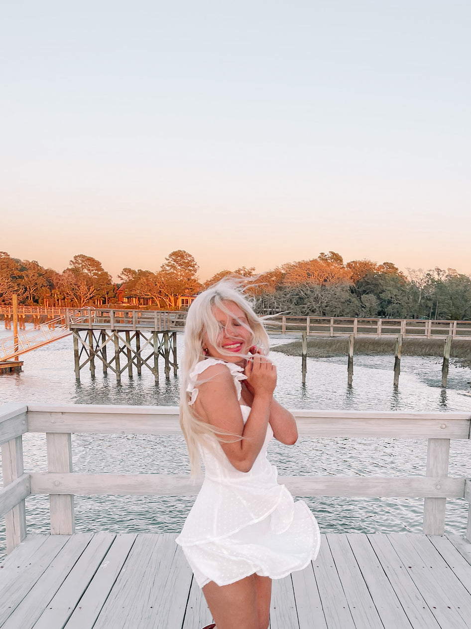 Woman in white summer dress on a dock overlooking calm water at sunset.
