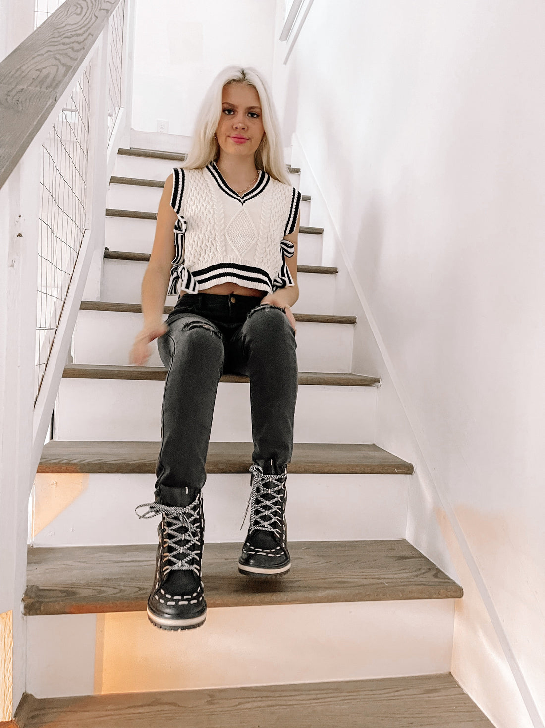 Woman in a cream cable-knit vest and black jeans sits on a staircase.
