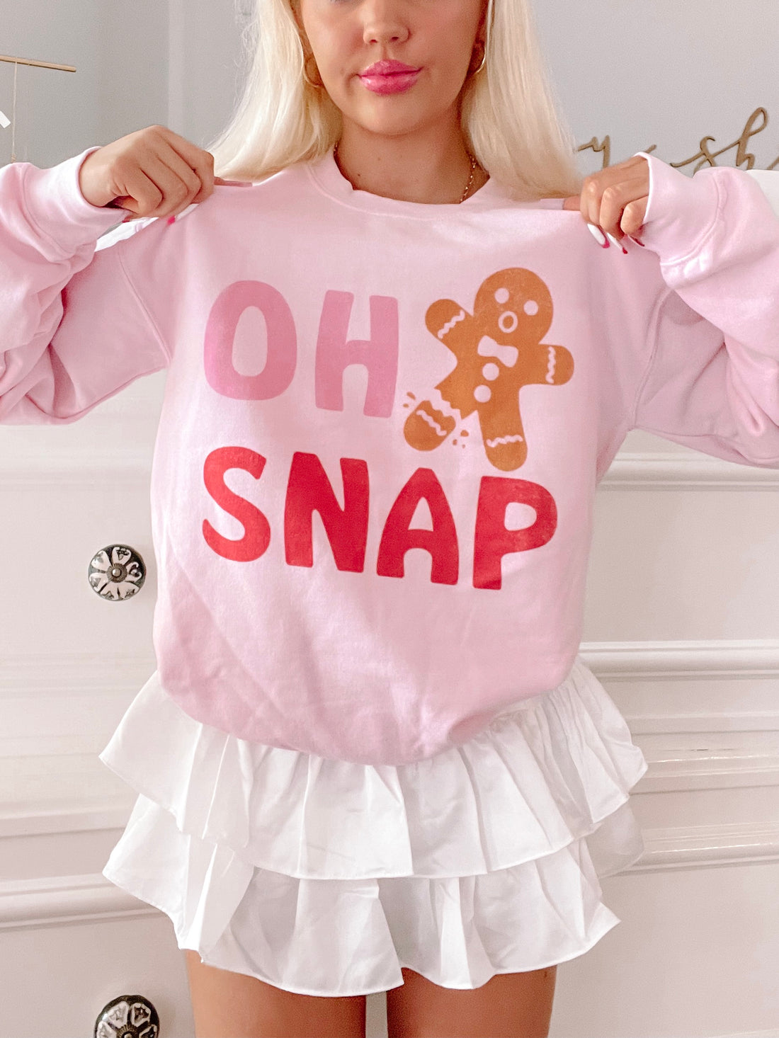 Pink crewneck sweatshirt with a whimsical gingerbread man design and "Oh Snap" text, modeled over a white layered skirt.
