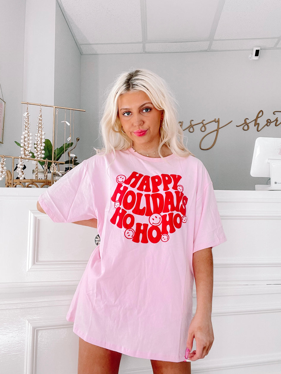 Pink oversized tee featuring "Happy Holidays Ho Ho Ho" in red lettering with smiley faces.
