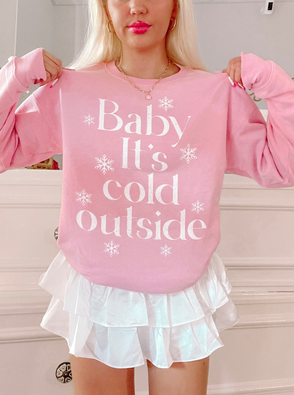 Pink crewneck sweatshirt with "Baby It's Cold Outside" text and snowflake accents, modeled with white skirt.

