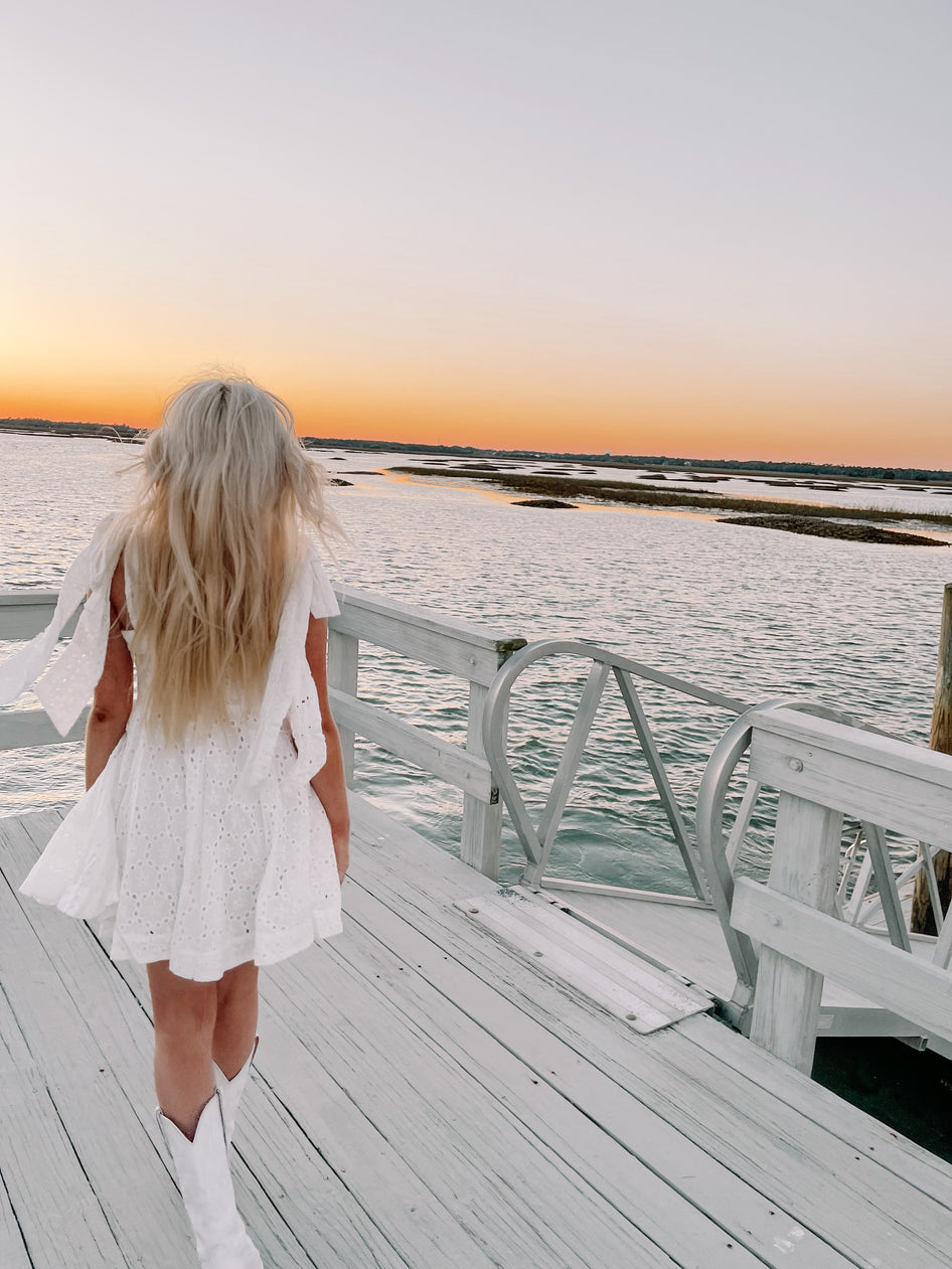 White eyelet dress on a pier overlooking sunset water views.
