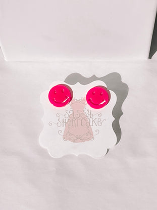 Bright pink smiley face earrings displayed on a white card.
