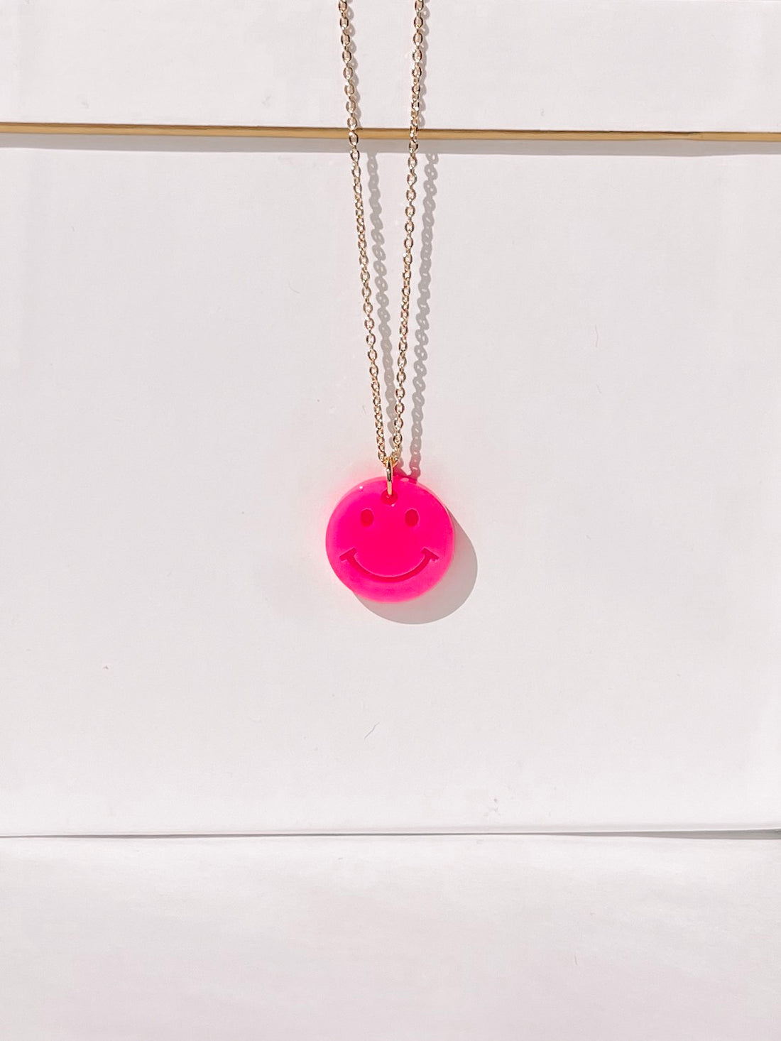Bright pink smiley face necklace on a gold chain against a white background.
