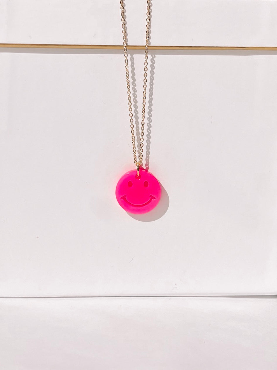 Bright pink smiley face necklace on a gold chain against a white background.
