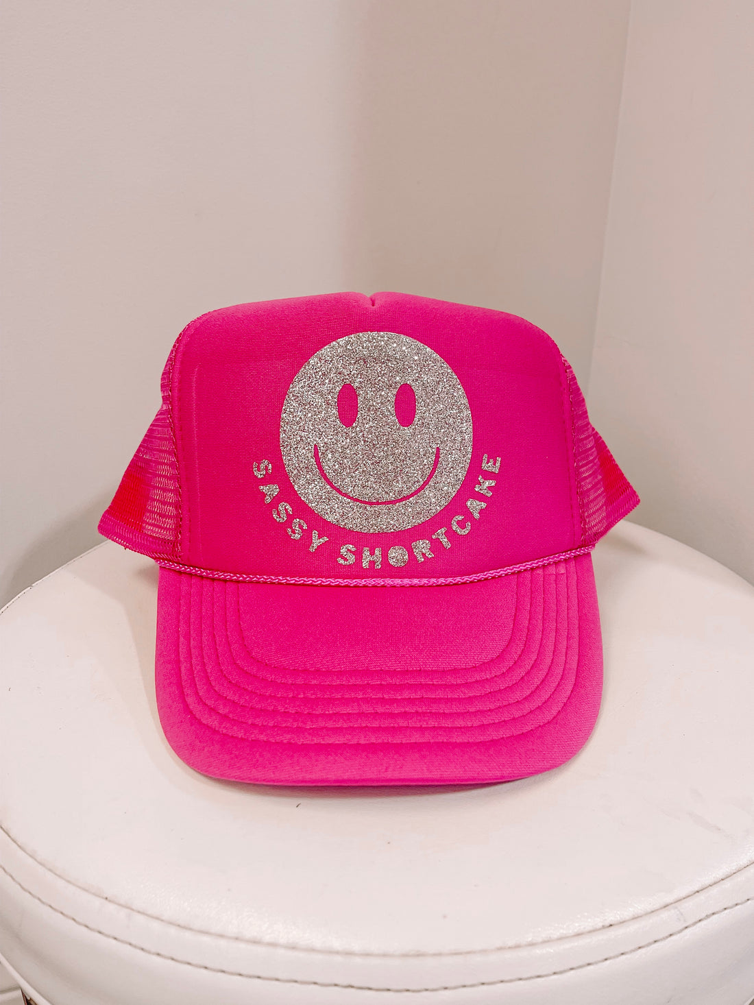 Hot pink trucker hat with a silver glitter smiley face and text detail, displayed on a white surface.
