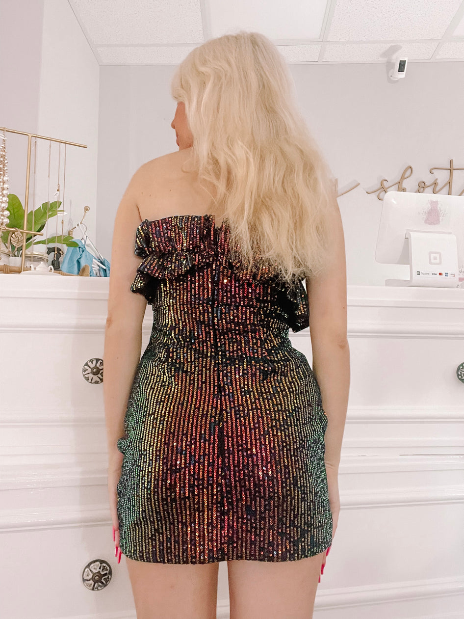 Sequined mini dress in iridescent rainbow hues, shown from the back against a white wall.
