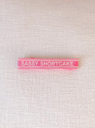 Pink woven bracelet label with white lettering on a white fabric background.
