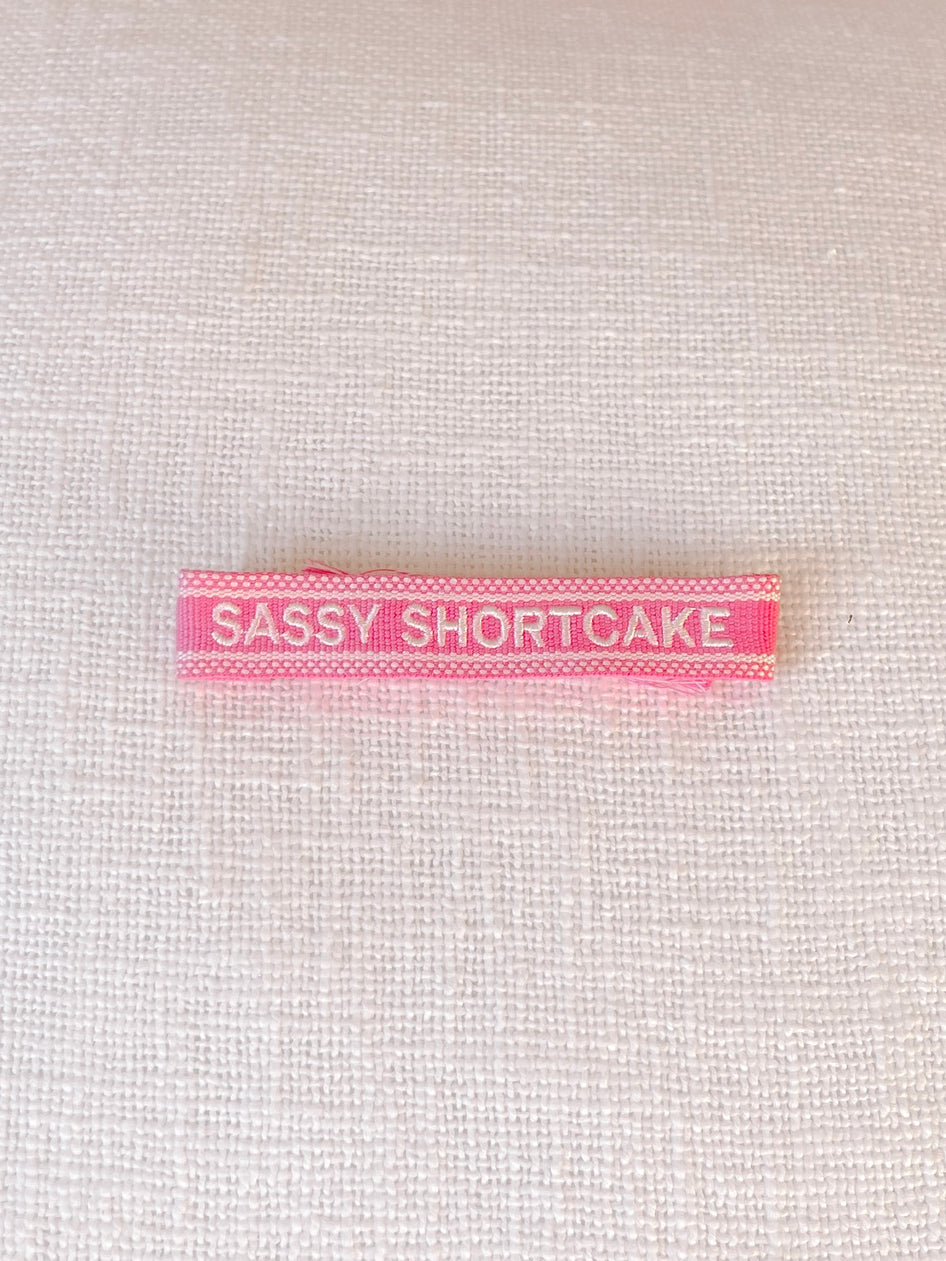 Pink woven bracelet label with white lettering on a white fabric background.
