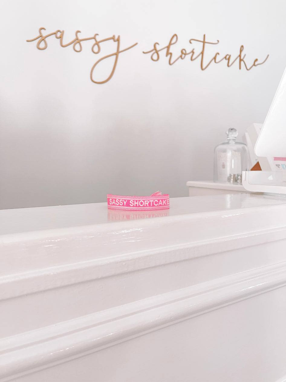 Pink ribbon bracelet with white lettering on a white counter.
