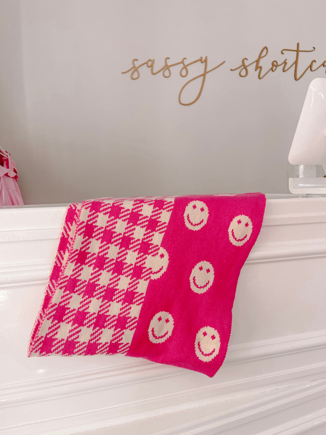 Pink and cream knitted scarf with a gingham pattern and smiley faces, displayed on a white surface.
