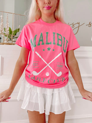 Hot pink golf tee featuring crossed golf clubs and "Est. 1986" detail, styled with a white ruffled skirt.
