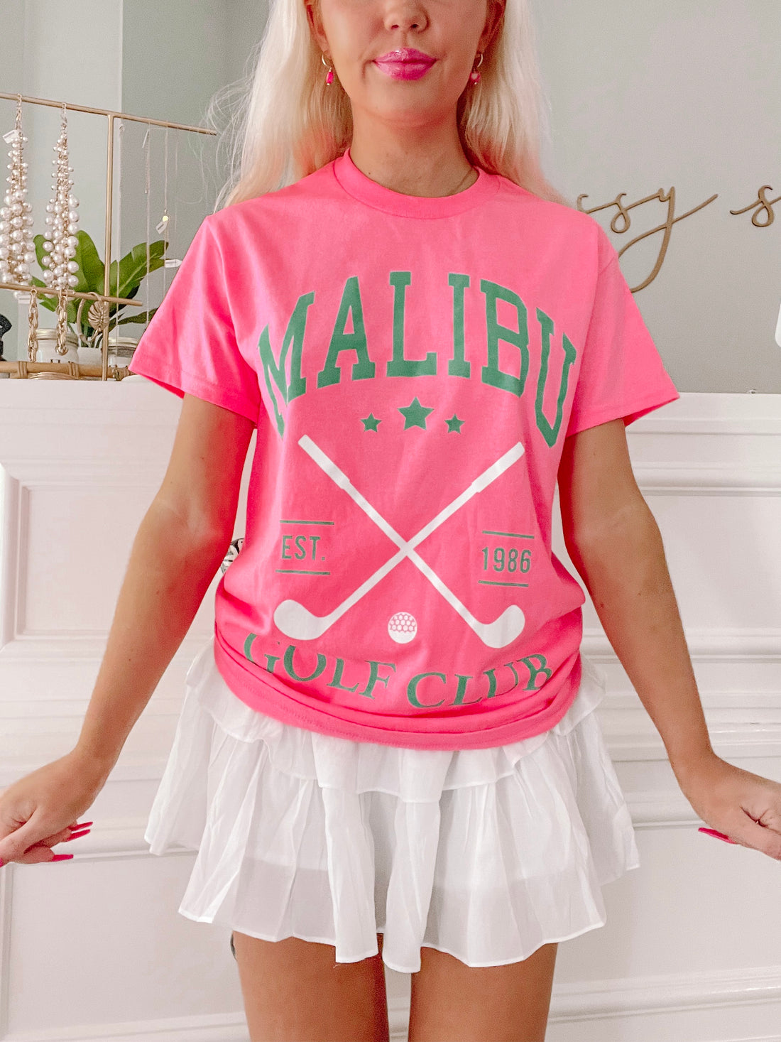 Hot pink golf tee featuring crossed golf clubs and "Est. 1986" detail, styled with a white ruffled skirt.
