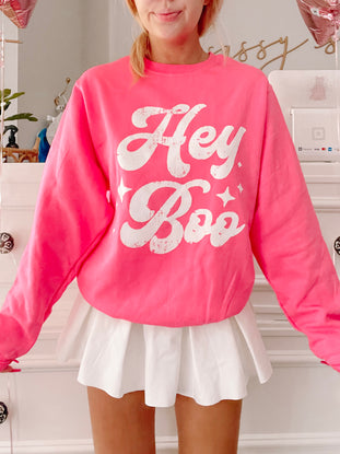 Hot pink crewneck sweatshirt with a retro "Hey Boo" graphic, styled with a white pleated mini skirt.