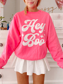Hot pink crewneck sweatshirt with a retro "Hey Boo" graphic, styled with a white pleated mini skirt.
