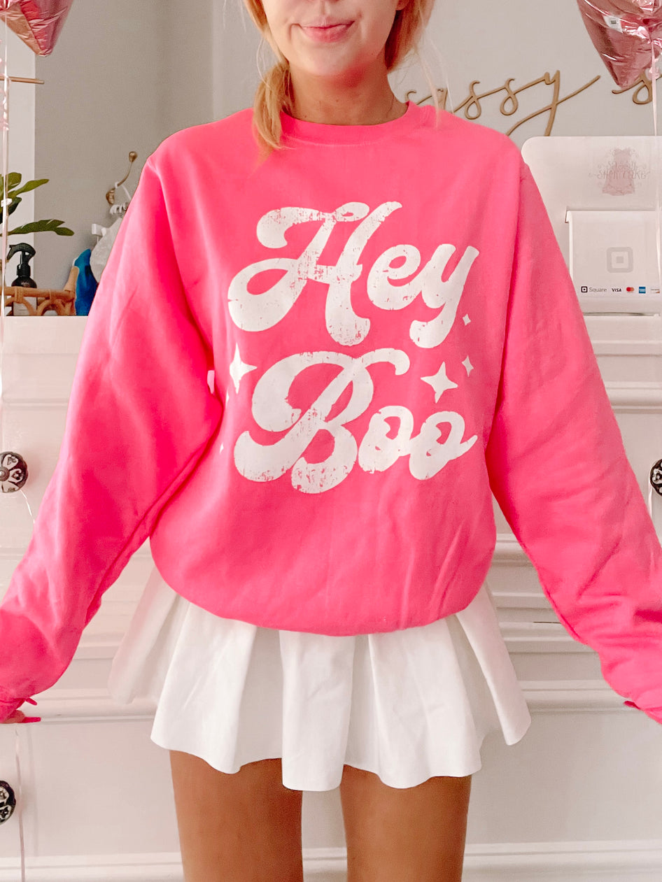 Hot pink crewneck sweatshirt with a retro "Hey Boo" graphic, styled with a white pleated mini skirt.
