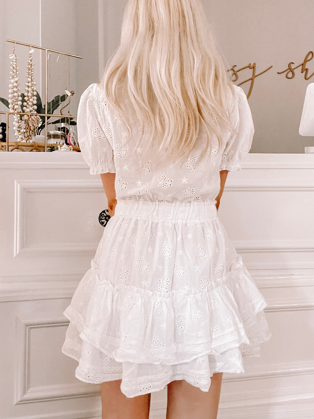 White eyelet mini dress with tiered ruffle skirt, shown from the back against a white wall.
