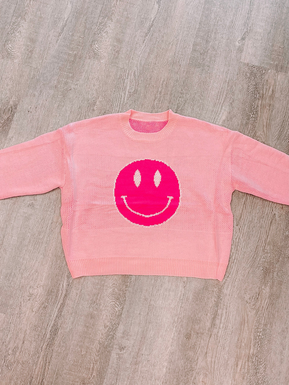Pink knit sweater featuring a bright pink smiley face, laid flat on a wood surface.
