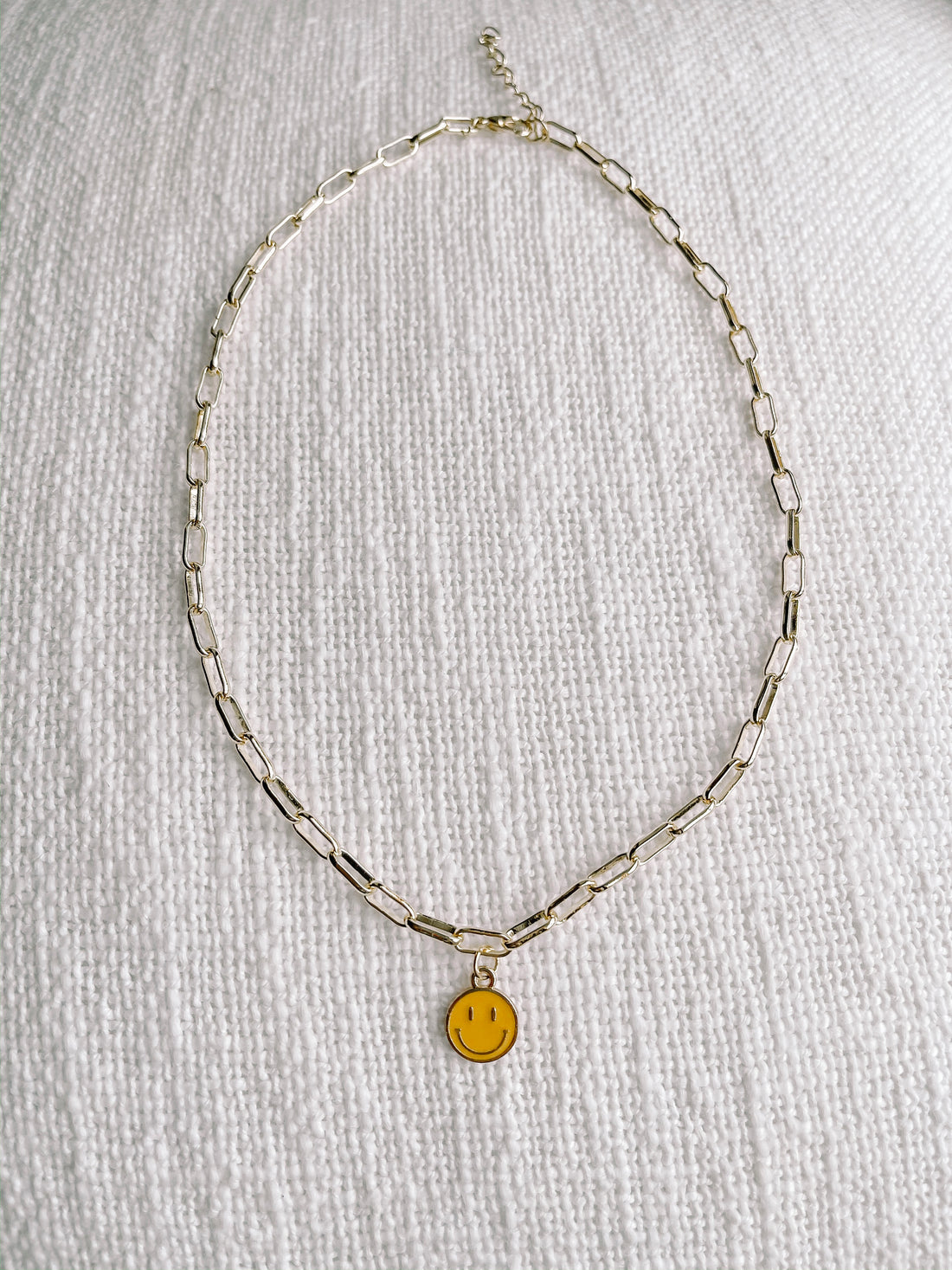 Gold chain necklace with cheerful yellow smiley face charm, displayed on white textured fabric.
