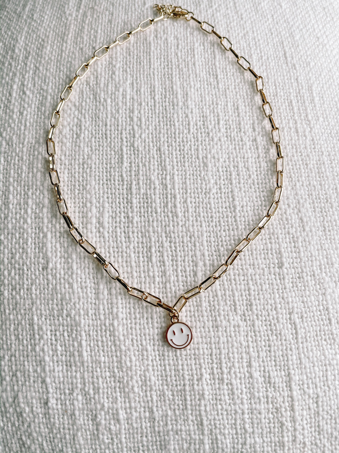 Gold chain necklace with a small white enamel smiley face charm, displayed on a textured white surface.
