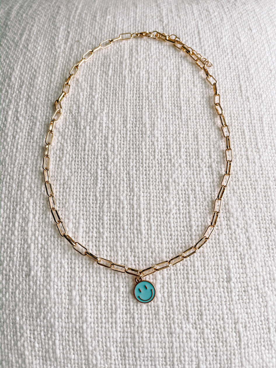 Gold chain necklace with light blue smiley face charm, displayed on textured white fabric.
