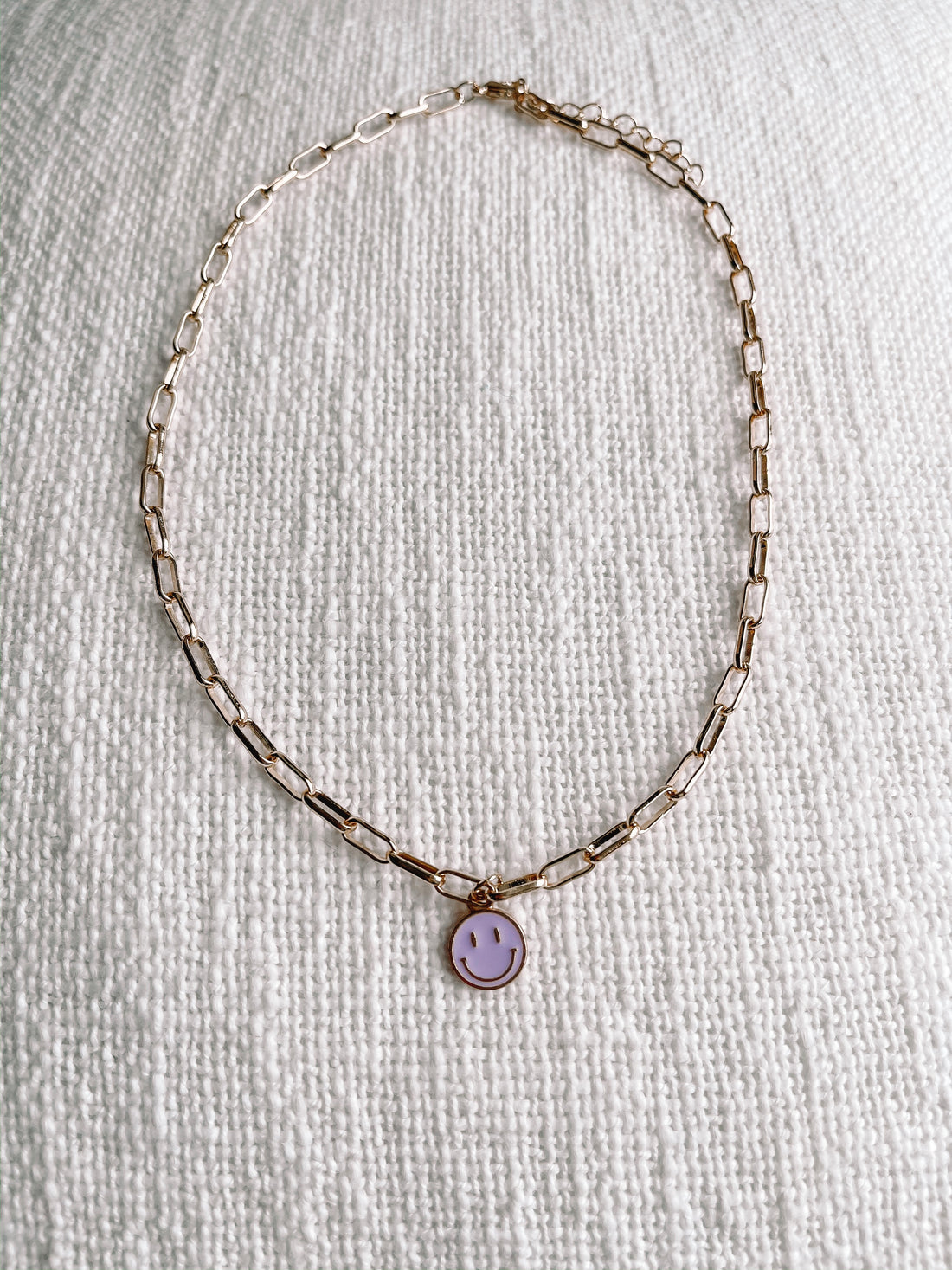 Gold chain necklace with lavender smiley face charm on textured white fabric
