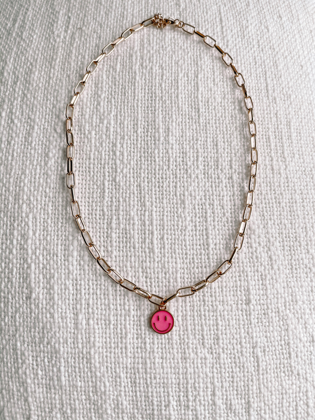 Gold chain necklace with a pink smiley face charm, displayed on a textured white surface.
