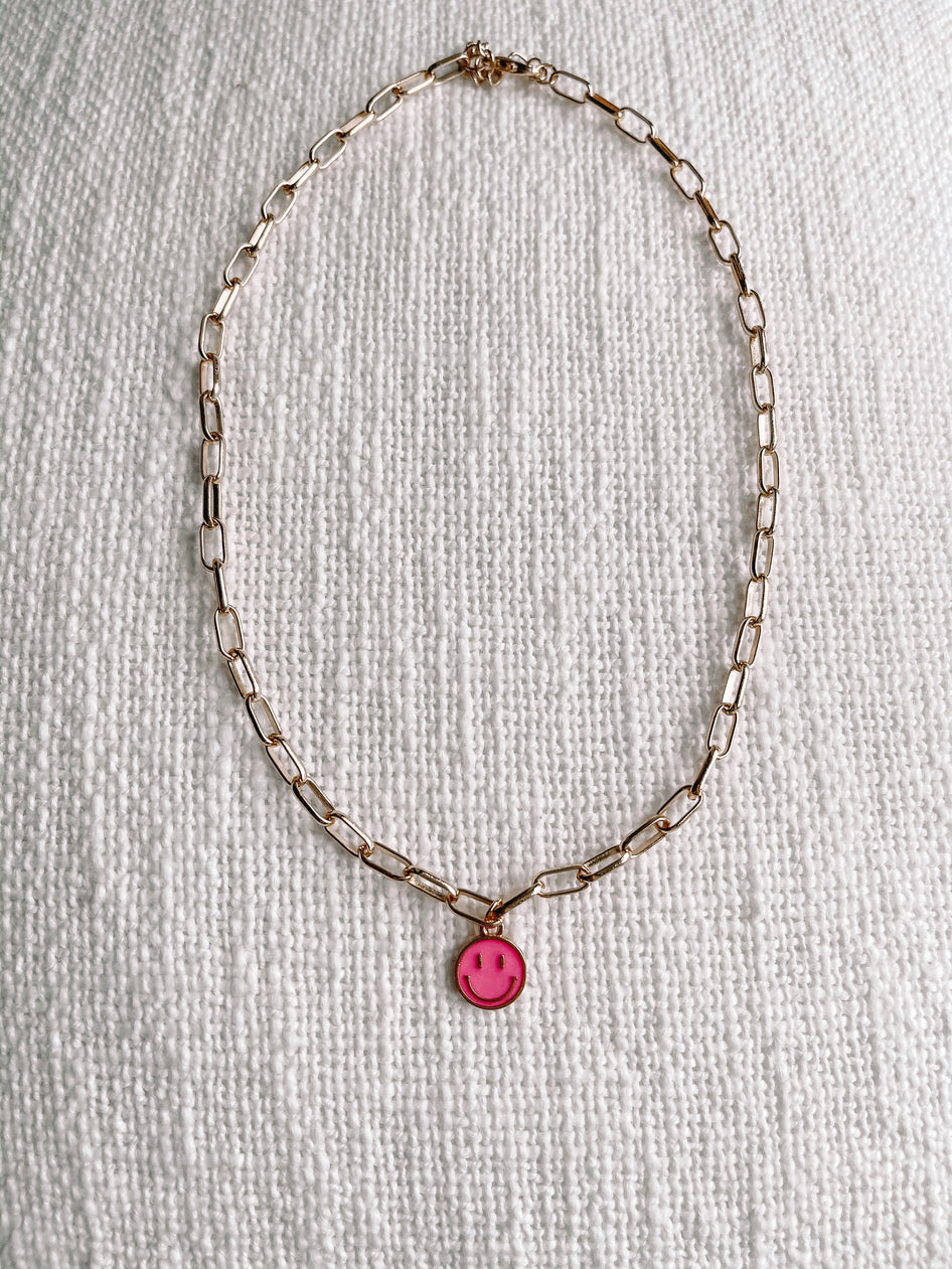 Gold chain necklace with a pink smiley face charm, displayed on a textured white surface.
