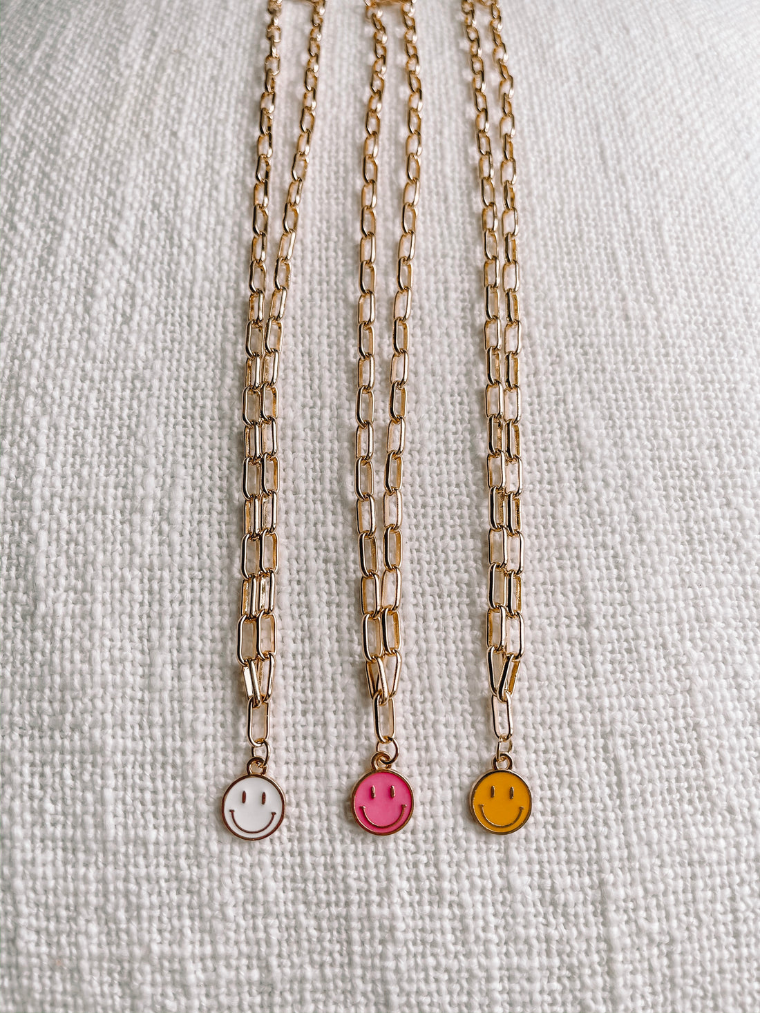 Gold chain necklaces with colorful smiley face charms on a textured white background.
