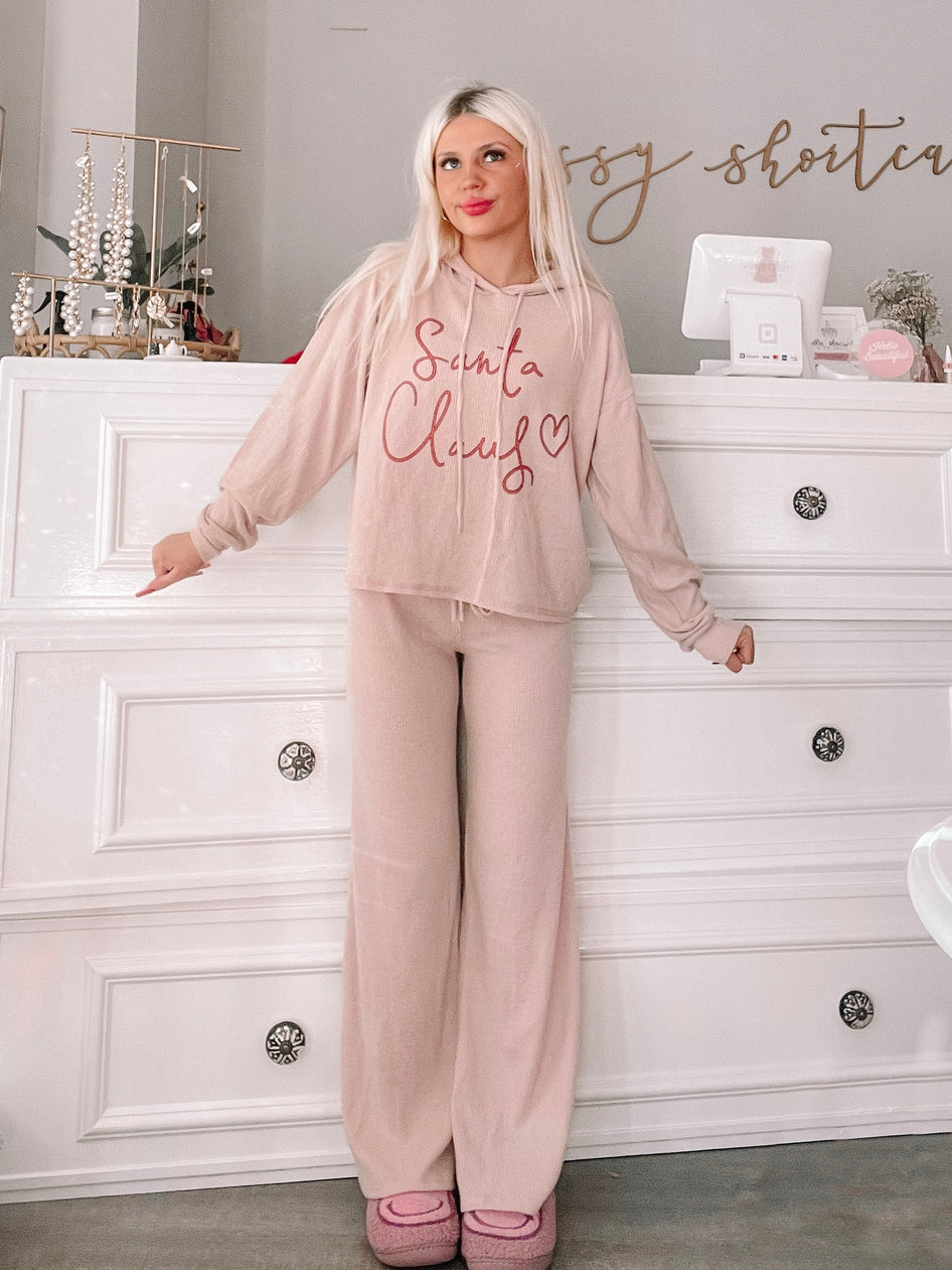 Cozy blush pink Christmas pajamas with a "Santa Claus" graphic, modeled against a white dresser.
